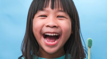 little girl smiles widely holding a toothbrush in front of a blue background